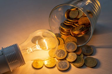 Ahşap yüzeyde, içi bozuk para dolu kavanozla birlikte parlak bir ampul yandı. Artan maliyetler. Tasarruf. Enerji tarifeleri.