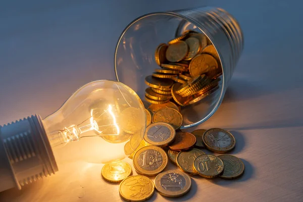 Incandescent light bulb turned on, with jar full of coins next to it, on wooden surface. Increased costs. Savings. Energy tariffs.