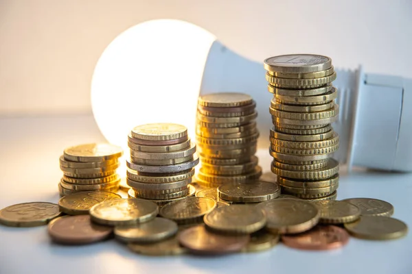 Stacks of euro coins with lit light bulb beside, placed on white wooden surface. Value of money and energy tariff trends.