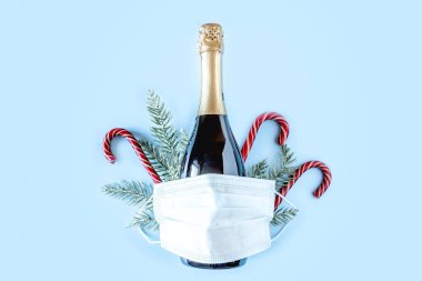 Şişe şampanya, ladin dalları, kırmızı şeker kamışı ve mavi arka planda koruyucu tıbbi maske. Koronavirüs enfeksiyonu salgını sırasında Noel ve Yeni Yıl. Düz yatıyordu.