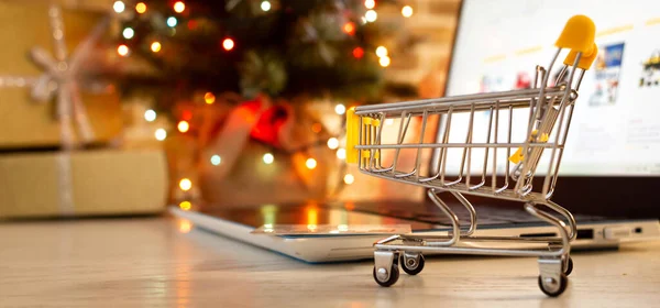 クリスマスオンラインショッピング 家でショッピングカートとノートパソコンぼやけたボケのライトとクリスマスツリーの背景に — ストック写真