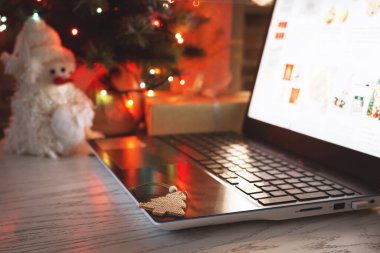 İnternetten Noel alışverişi. Evdeki masadaki dizüstü bilgisayar, bulanık ışık saçan bir Noel ağacının arka planında..