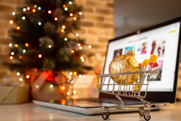 İnternetten Noel alışverişi. Evde dizüstü bilgisayar ve alışveriş arabası bulanık ışıkları olan bir Noel ağacının arka planında..