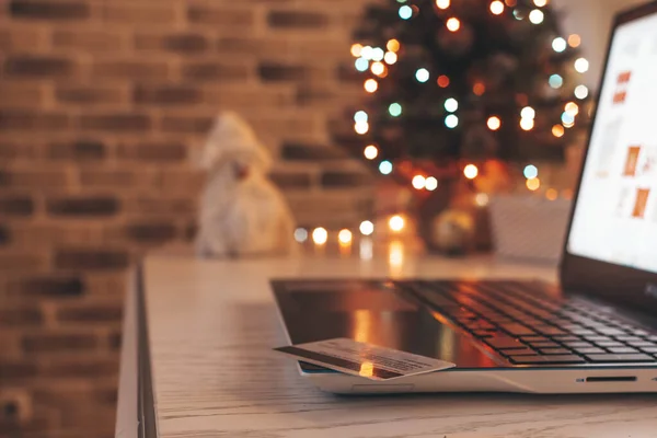İnternetten Noel alışverişi. Laptop ve kredi kartı evdeki masanın üzerinde bulanık ışıklı bir Noel ağacının arka planında..