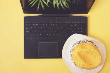 Laptop, hasır şapka ve sarı arka planda palmiye yaprakları. Yaz tatili, tatil, online rezervasyon, planlama seyahat konsepti. Düz yatış, üst görünüm.