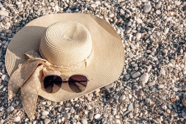 Women's straw hat with a bow and sunglasses on background of a pebble beach. Summer holiday, vacation and travel concept. Copy space