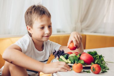 Çocuk mutfakta oturmuş sağlıklı sebze, salatalık ve domates yiyor.