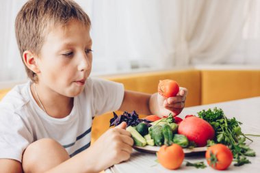 Çocuk mutfakta oturmuş sağlıklı sebze, salatalık ve domates yiyor.