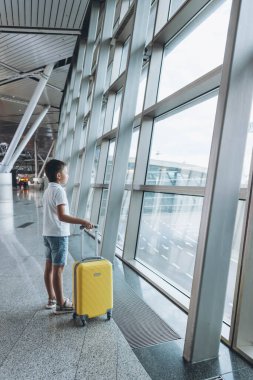 Boş havaalanı terminalinde sarı valizli bir çocuk pencereden dışarı bakıyor. Tişörtlü ve şortlu çocuk salonda uçak uçuşunu bekliyor. Aile gezisi konsepti