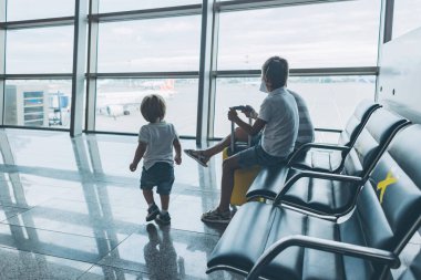 Boş havaalanı terminalindeki çocuklar pencereden dışarı bakıyorlar. Koruyucu tıbbi maske takan küçük çocuklar salonda oturmuş uçak uçuşunu bekliyorlar. Aile gezisi konsepti