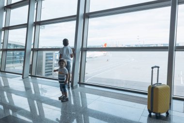 Boş havaalanında sarı valizli iki erkek kardeş. Çocuklar pencereden uçağa bakıyor. Çocuklarla seyahat ve uçmak. Aile çıkış kapısını bekliyor. Aile gezisi ve tatil konsepti.