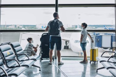 Baba ve oğulları boş havaalanında sarı valizli koruyucu tıbbi maske takıyor. Çocuklar pencereden uçağa bakıyor. Aile çıkış kapısını bekliyor. Aile gezisi ve tatil konsepti.