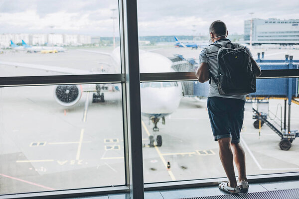 человек в защитной медицинской маске с рюкзаком, стоящим в пустом аэропорту и ожидающим отъезда. Пассажир в дерьме смотрит в окно на самолет. путешествие во время пандемии коронавируса