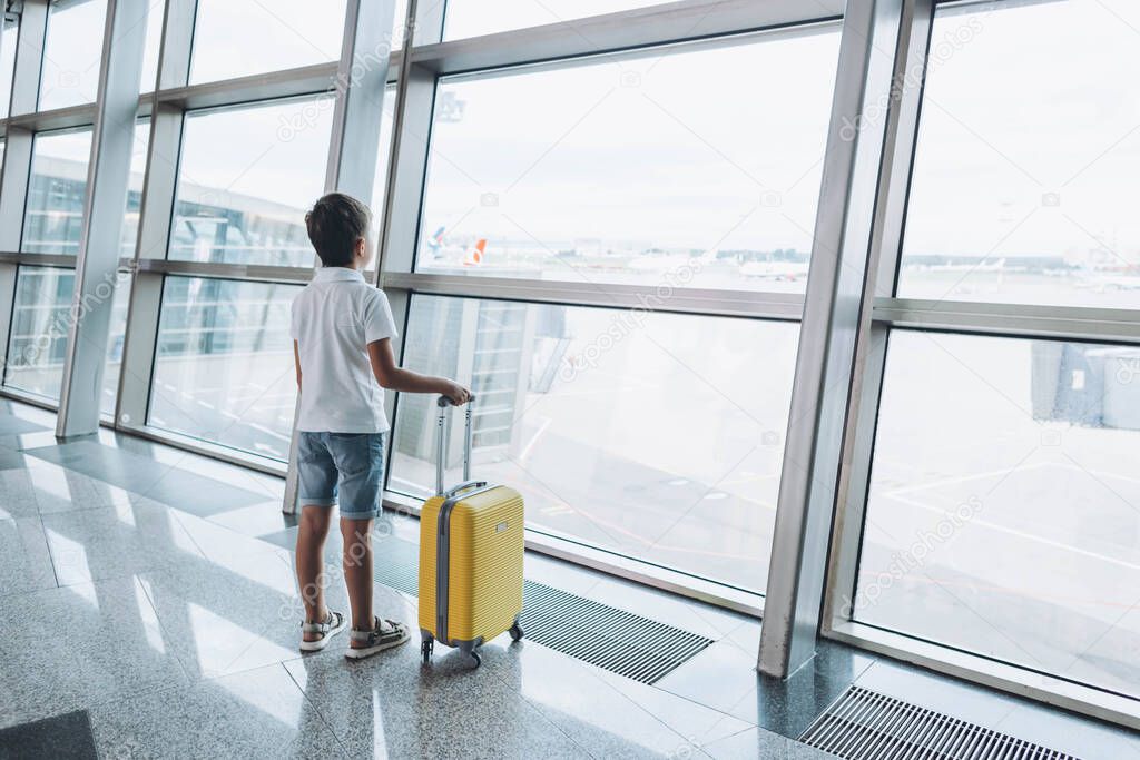 Boy with yellow suitcase at empty airport terminal waiting for departure looking out the window. Child in T-shirt and shorts stands at lounge waiting for plane flight. Family trip concept