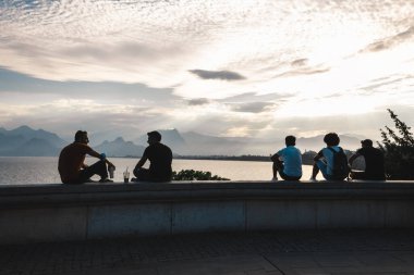 Antalya, Türkiye-Temmuz 2021: Bir grup genç gezgin taştan korkuluklara oturmuş denize bakıyor. Türkiye 'nin Antalya kentinde dağların ve denizin tadını çıkaran turistler. Etkin yaşam tarzı kavramı