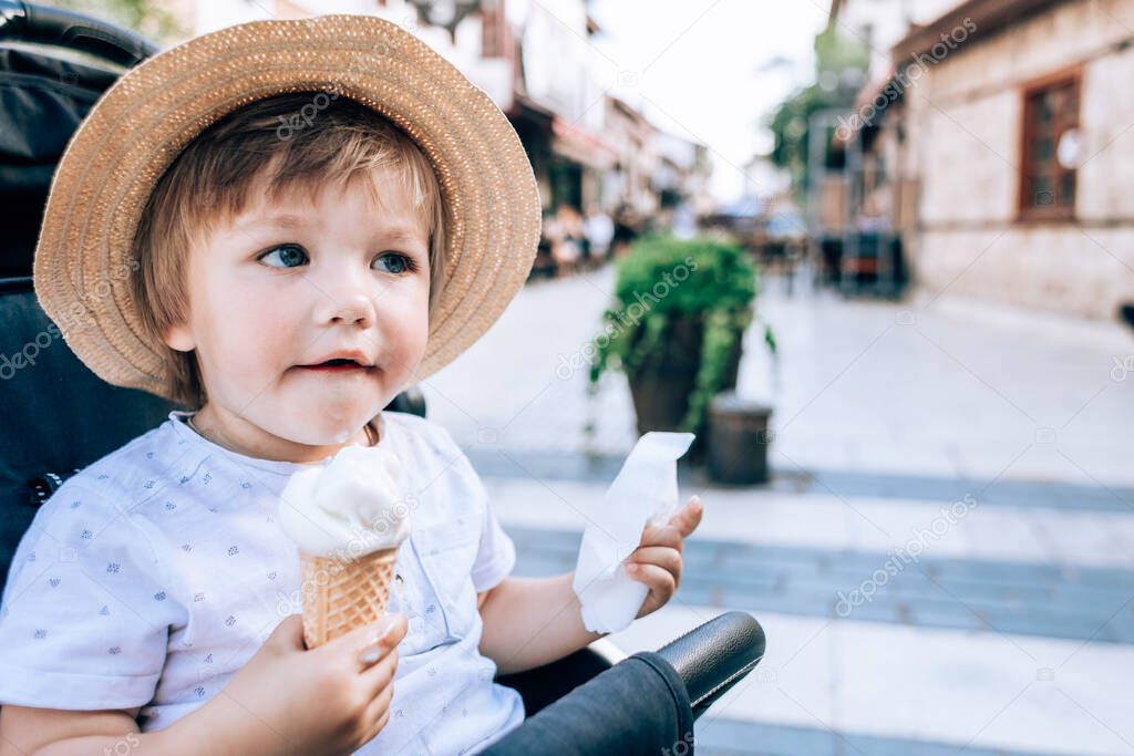 Portrait of cute little boy in straw hat sitting in stroller with ice cream. Toddler eating ice cream in a waffle cone on cityscape background. Dairy free ice cream.