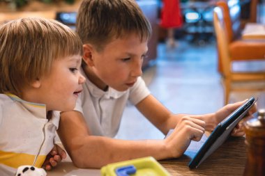Çocuk ve bebek, restoranda siparişleri beklerken internetten tablette video oyunu oynuyorlar. Akıllı telefon kullanan çocuklar. Çocuklar teknolojiyi kullanıyor. Ekran zaman bağımlılığı.