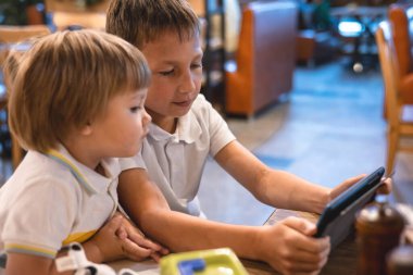 Çocuk ve bebek, restoranda siparişleri beklerken internetten tablette video oyunu oynuyorlar. Akıllı telefon kullanan çocuklar. Çocuklar teknolojiyi kullanıyor. Ekran zaman bağımlılığı.
