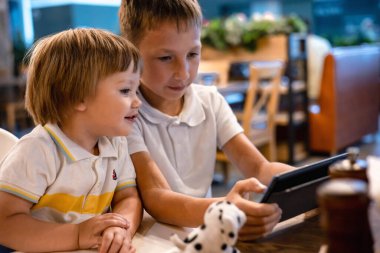 Çocuk ve bebek, restoranda siparişleri beklerken internetten tablette video oyunu oynuyorlar. Akıllı telefon kullanan çocuklar. Çocuklar teknolojiyi kullanıyor. Ekran zaman bağımlılığı.