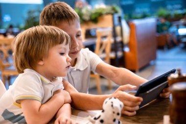 Çocuk ve bebek, restoranda siparişleri beklerken internetten tablette video oyunu oynuyorlar. Akıllı telefon kullanan çocuklar. Çocuklar teknolojiyi kullanıyor. Ekran zaman bağımlılığı.
