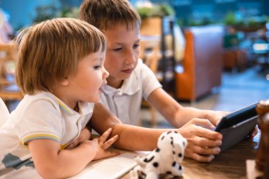 Çocuk ve bebek, restoranda siparişleri beklerken internetten tablette video oyunu oynuyorlar. Akıllı telefon kullanan çocuklar. Çocuklar teknolojiyi kullanıyor. Ekran zaman bağımlılığı.