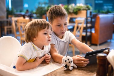 Çocuk ve bebek, restoranda siparişleri beklerken internetten tablette video oyunu oynuyorlar. Akıllı telefon kullanan çocuklar. Çocuklar teknolojiyi kullanıyor. Ekran zaman bağımlılığı.