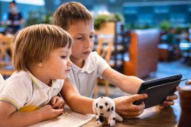 Çocuk ve bebek, restoranda siparişleri beklerken internetten tablette video oyunu oynuyorlar. Akıllı telefon kullanan çocuklar. Çocuklar teknolojiyi kullanıyor. Ekran zaman bağımlılığı.