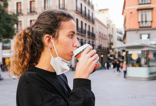 New normal. Smiling teenager drinking coffee on the street during the pandemic. Smiley young girl drinking coffee at Christmas. Drinking a coffee on the street with a mask. Christmas. Hot coffee.