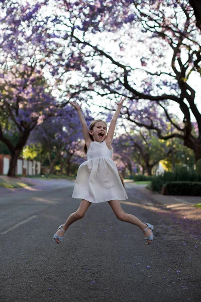 白いドレスを着た長髪のかわいい女の子がプレトリアのジャカランダの木を咲かせて通りをジャンプします — ストック写真
