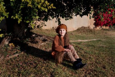 Küçük bir kız, kahverengi elbiseli, uzun saçlı, büyük köklü bir ağacın yanında oturuyor. Sonbahar portresi. Küçük Fransız kadın. Güney Afrika 'da kış.