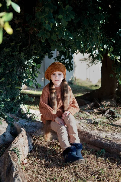 Little girl, schoolgirl in brown clothes with long hair, sitting near tree with large roots. Autumn portrait. Little Frenchwoman. Winter in South Africa.