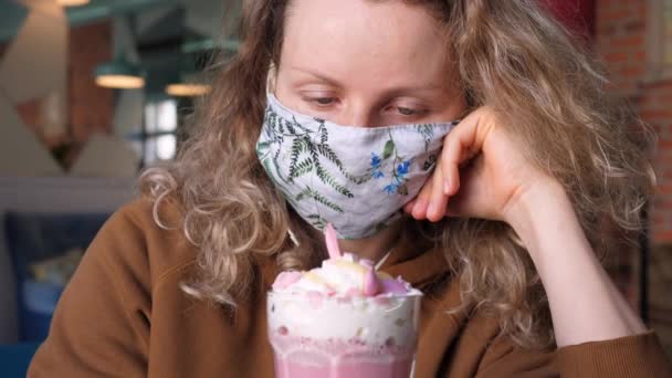 Женщина в защитной маске для лица в кафе смотрит на розовый кофе латте с взбитыми сливками — стоковое видео