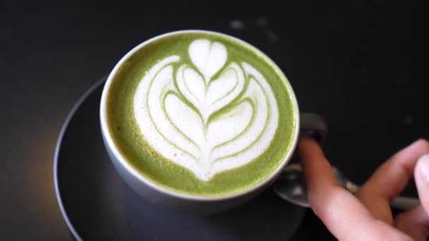 Top view of a hand picking up black cup of matcha tea latte with latte art on it 