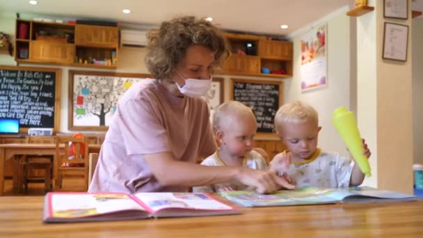 Молодая белая мать в маске читает книги с дочерьми-близнецами в детской. — стоковое видео