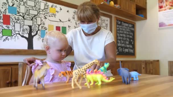 La abuela con mascarilla le enseña a su nieta acerca de los dinosaurios y otros animales con juguetes de colores. Tiempo de vinculación de calidad — Vídeo de stock