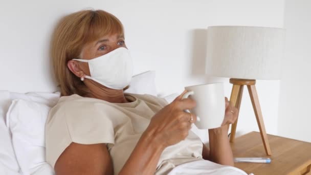 Vista lateral de una mujer caucásica de mediana edad con una máscara protectora en su cama sosteniendo una taza blanca mirando al espacio — Vídeo de stock