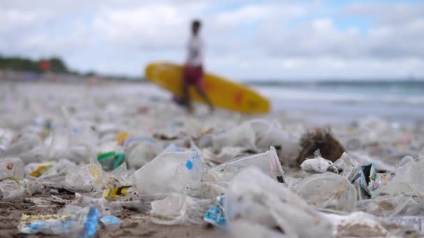 Sluiten van plastic vervuiling op het strand. Man met surfplank op de achtergrond — Stockvideo