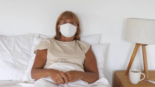 Retrato de senhora caucasiana doente em máscara protetora deitada em sua cama olhando para a câmera — Vídeo de Stock