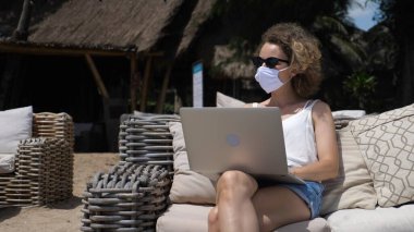 Dijital göçebe yaşam tarzı konsepti. Koruyucu yüz maskeli beyaz kız sahildeki kanepede bilgisayarla çalışıyor.
