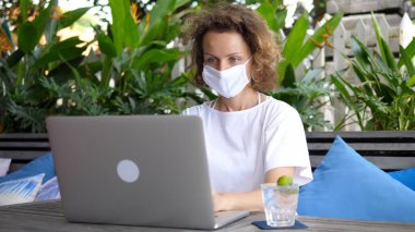  Uzaktan çalışma konsepti. Koruyucu yüz maskesi takan beyaz kadın dışarıda dizüstü bilgisayarda çalışıyor. Arkaplanda Tropik Yapraklar.