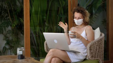Koruyucu maskeli genç bir kadın avuçlarıyla terasta video görüşmesinin keyfini çıkarıyor. Tatili boyunca sevdiklerine bağlı kalmak.