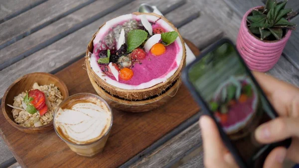 Bovenaanzicht van veganistisch ontbijt van smoothie kokosnoot kom geserveerd op houten bord met zuivel gratis latte wordt gefotografeerd met een smartphone — Stockfoto