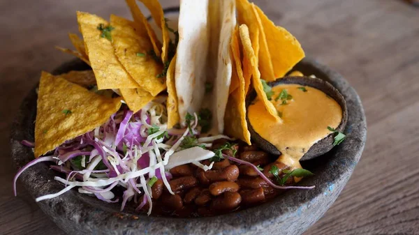 Close up of hand spinning stone bowl with vegan snacks in mexican style: nachos, red kidney beans, guacamole and sauce. Vegan street food