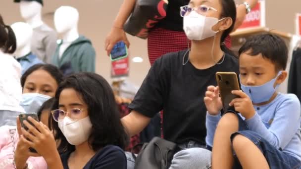 Azjatyckie dzieci w maskach, siedzące na schodach i wpatrujące się w smartfony w centrum handlowym. Bali-grudzień-2020 — Wideo stockowe