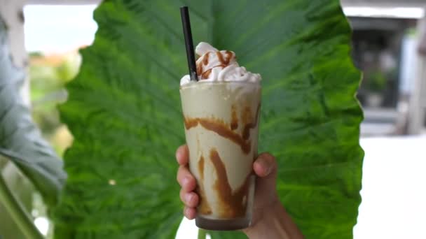 Hand tonen aan de camera glas van iced matcha groene thee met slagroom op de achtergrond van een gigantisch blad — Stockvideo