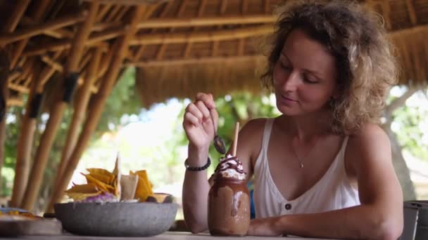 La rubia soñadora disfruta de la crema batida de su batido durante el almuerzo en un bar al aire libre de la playa. Estilo de vida tropical — Vídeos de Stock