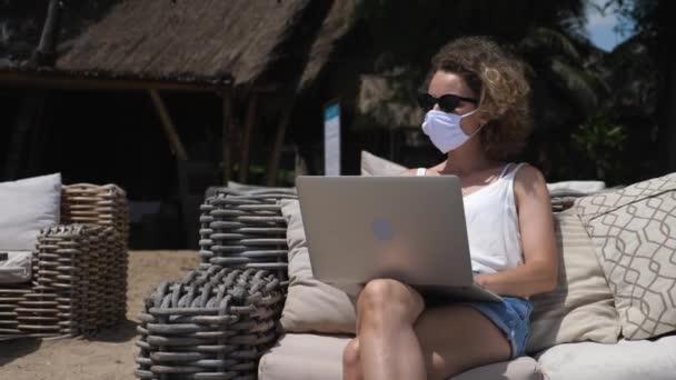 Концепція цифрового кочового способу життя. Кавказька дівчина в захисній масці працює на комп'ютері прямо з дивана на пляжі. — стокове відео