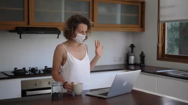 Caucasian girl in face mask video chatting and drinking tea at the kitchen. Staying connected during quarantine — Stock Video