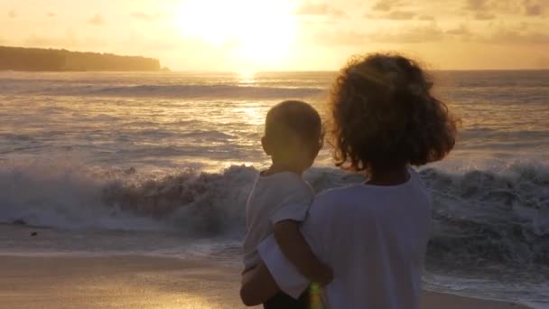 Achteraanzicht van een vrouw die een baby vasthoudt en de oceaan bekijkt bij zonsondergang. Moeder kind speciale band — Stockvideo