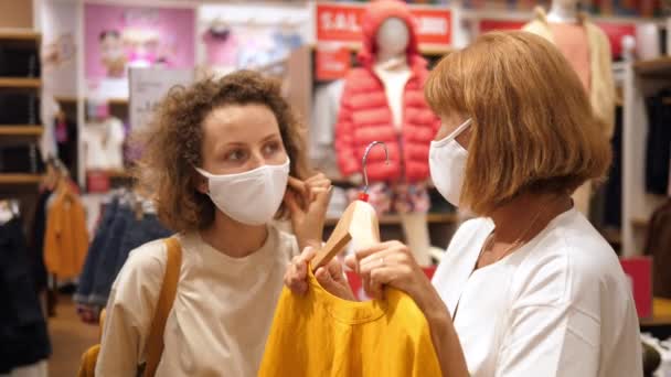 Señoras jóvenes y de mediana edad en máscaras faciales discutiendo algo mientras compran ropa — Vídeo de stock